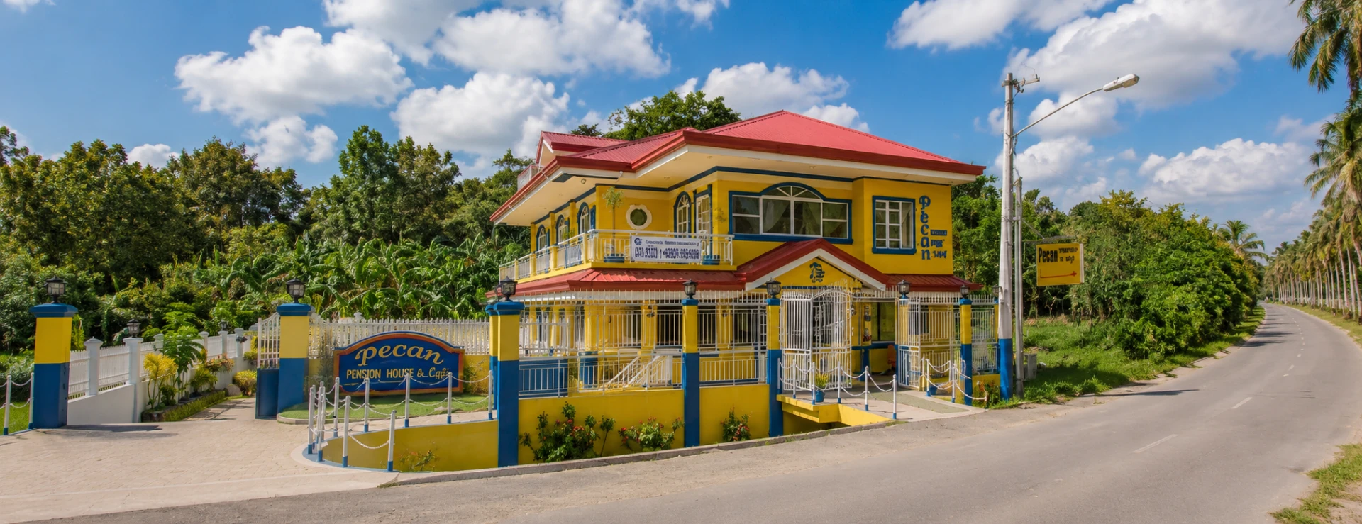 Panoramic view of Pecan Pension House & Café and its surroundings, showing the bright yellow and blue building, entrance gate, Pecan signage, and nearby road near Iloilo International Airport